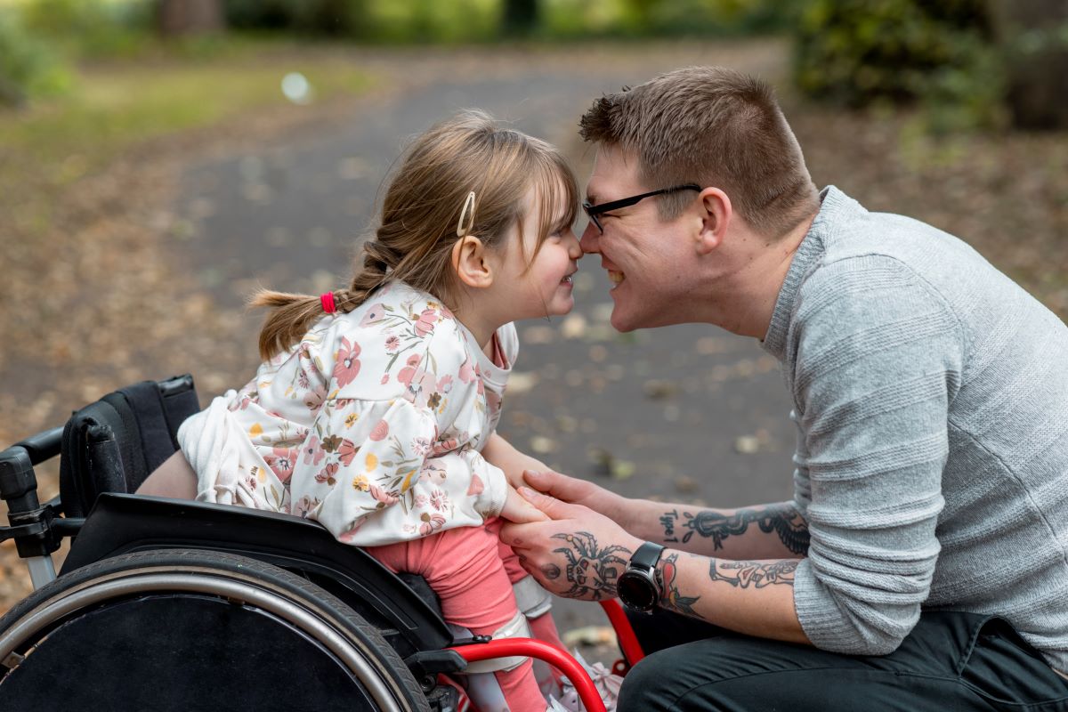 Older person in a park is crouched in order to press their nose against the nose of a child in a wheelchair opposite of them. Both people are smiling.