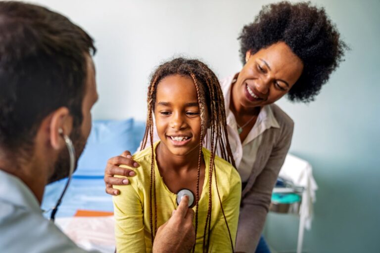 Mother behind daughter holding daughters shoulder as a doctor places a listening device on a child's chest in order to hear their lungs