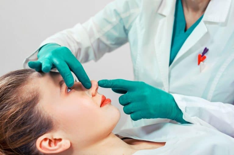 Image starts at just below chest height. Woman is laying down in medical outfit. The upper body and arms of a doctor wearing scrubs is connected to gloved hands which are placing a piece of medical tape onto the womans nose as she looks up at them from the table.