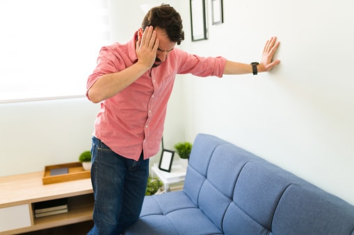 Man in pink shirt in an office setting holding his head with one hand while propping himself up against a wall with his other arm outstretched.