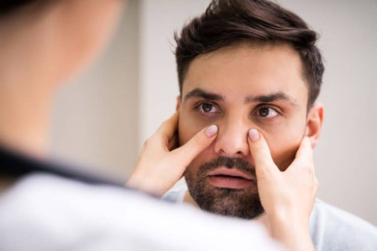 Blurry back shoulder and face of a person pressing their thumbs on either side of the bridge of the nose of a man who is looking at them.