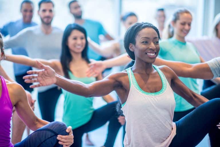 Focus on a woman in a floor class who is doing stretches with women and men. Everyone is balancing on one leg. The other leg is held up with one hand by the knee. The opposite arm on the standing leg is held outward and horizontally.