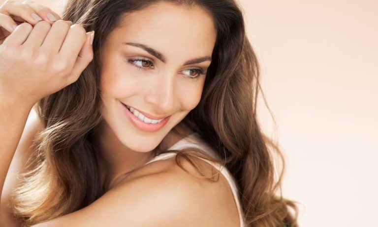 Closeup of brunette woman with hazel eyes smiling and looking towards the right while touching her hair. The background is beige.