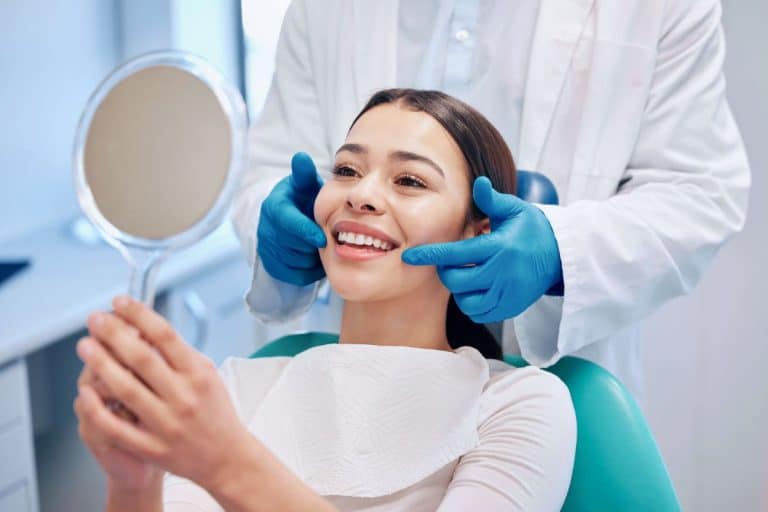 A woman in a dental style chair looks in a mirror and smiles. A doctor is behind her using gloved hands to point at her checks with gloved hands.