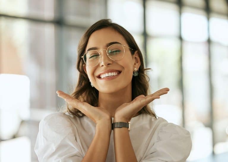 Woman wearing glasses is smiling with her eyes shut. The photo is from the shoulder up. She holds her hands out and below her chin in the shape of an open book. The background is blurry but indicates an open office setting.