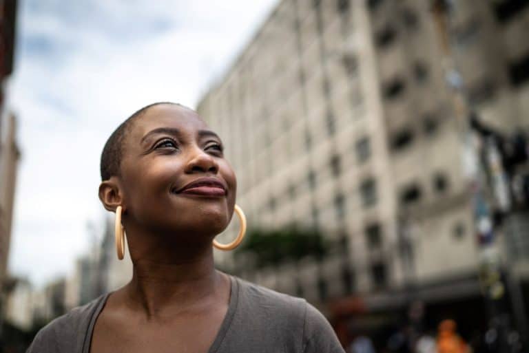 A woman on a city street. Image starts at shoulder height. She is wearing large gold hoop earrings and looking off frame to the upper right. She is smiling with a closed mouth and appears content.